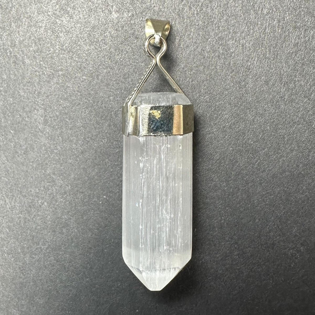 Clear selenite pendant with a pointed, elongated crystal held by a geometric sterling silver clasp on a textured gray background.