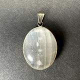 Oval selenite pendant with translucent white striations set in a simple sterling silver clasp on a dark gray background.