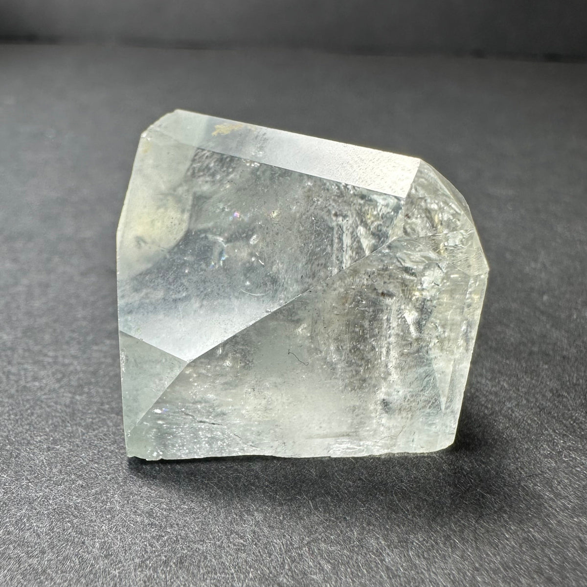 Clear and slightly cloudy white topaz crystal with natural faceted surfaces resting on a dark gray background, showing internal textures and light reflections.