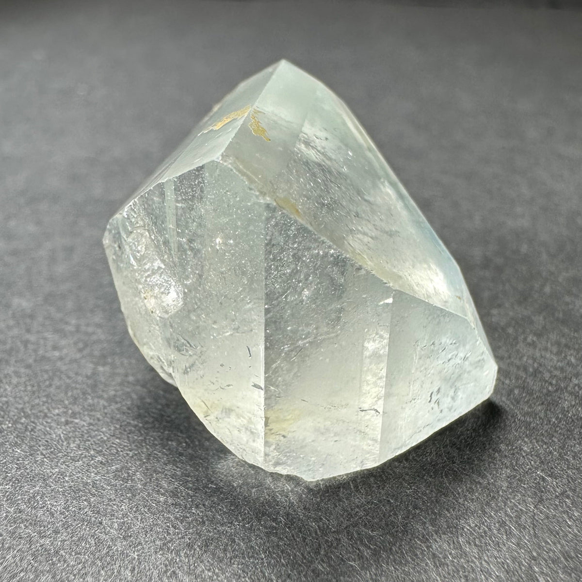 Translucent white topaz crystal with natural faceted surfaces and subtle internal inclusions, resting on a dark gray textured surface.