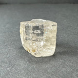 Transparent golden topaz crystal with subtle internal fractures, resting on a dark gray textured surface, showing a roughly cube-shaped form with a slightly irregular top.