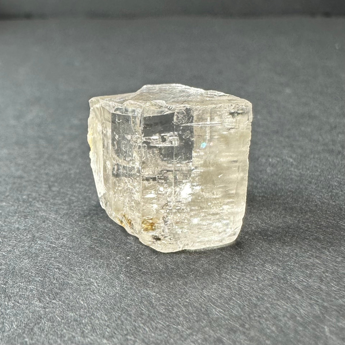 Transparent golden topaz crystal with subtle internal fractures, resting on a dark gray textured surface, showing a roughly cube-shaped form with a slightly irregular top.