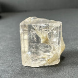 Transparent golden topaz crystal with irregular cubic shape and internal fractures, resting on a gray surface with a slightly blurred background