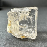 Transparent golden topaz crystal with rough edges and internal fractures, resting on a dark textured surface, showing natural inclusions of pale yellow color.