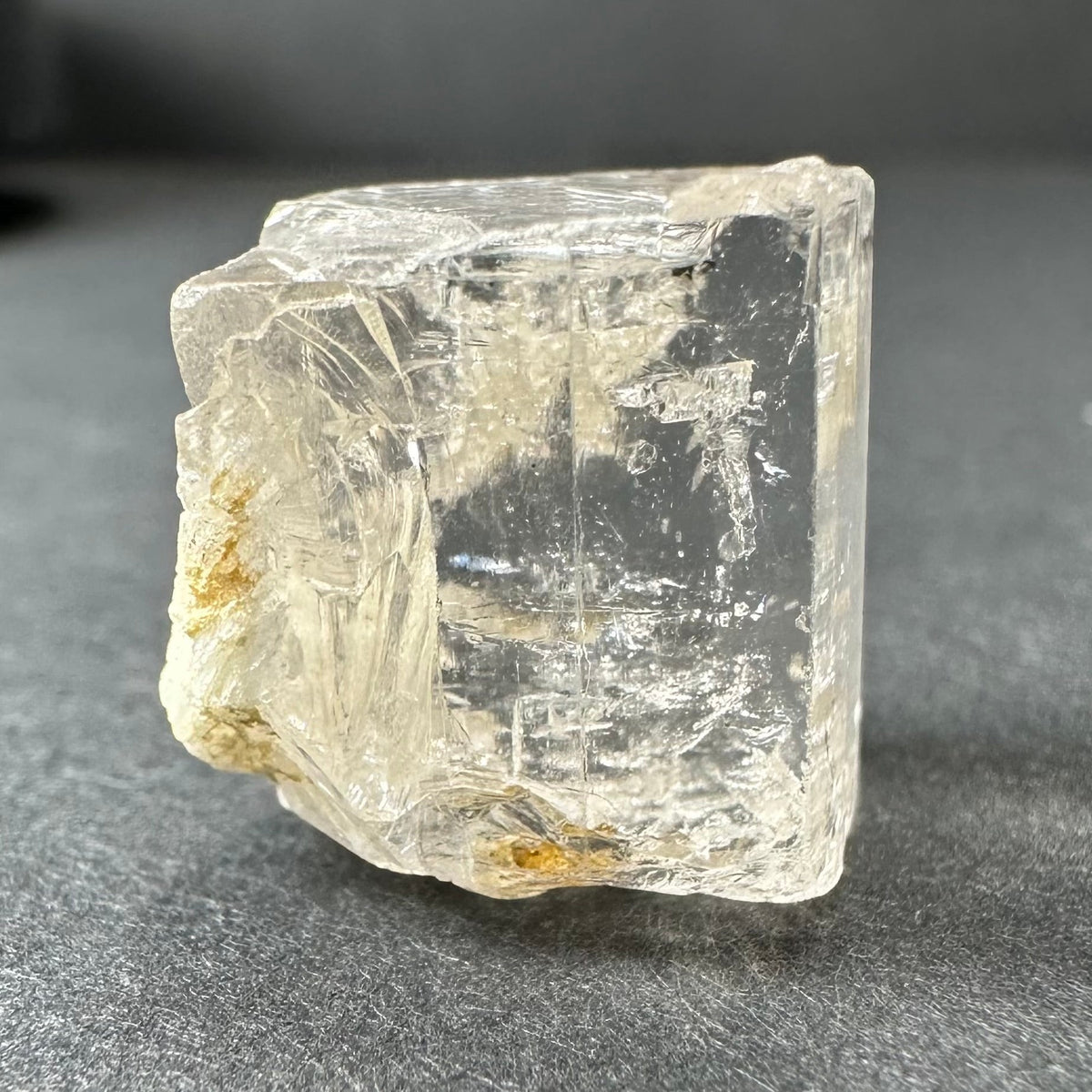 Transparent golden topaz crystal with rough edges and internal fractures, resting on a dark textured surface, showing natural inclusions of pale yellow color.
