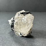 Transparent white topaz crystal with embedded black tourmaline strands, resting on a gray surface with a dark gray background.