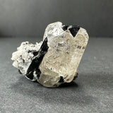 Transparent white topaz crystal with sharp black tourmaline inclusions resting on a dark gray surface with a neutral gray background.