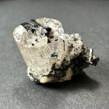 Clear white topaz crystal cluster with scattered glossy black tourmaline crystals on a gray surface, highlighting contrasting transparency and texture.