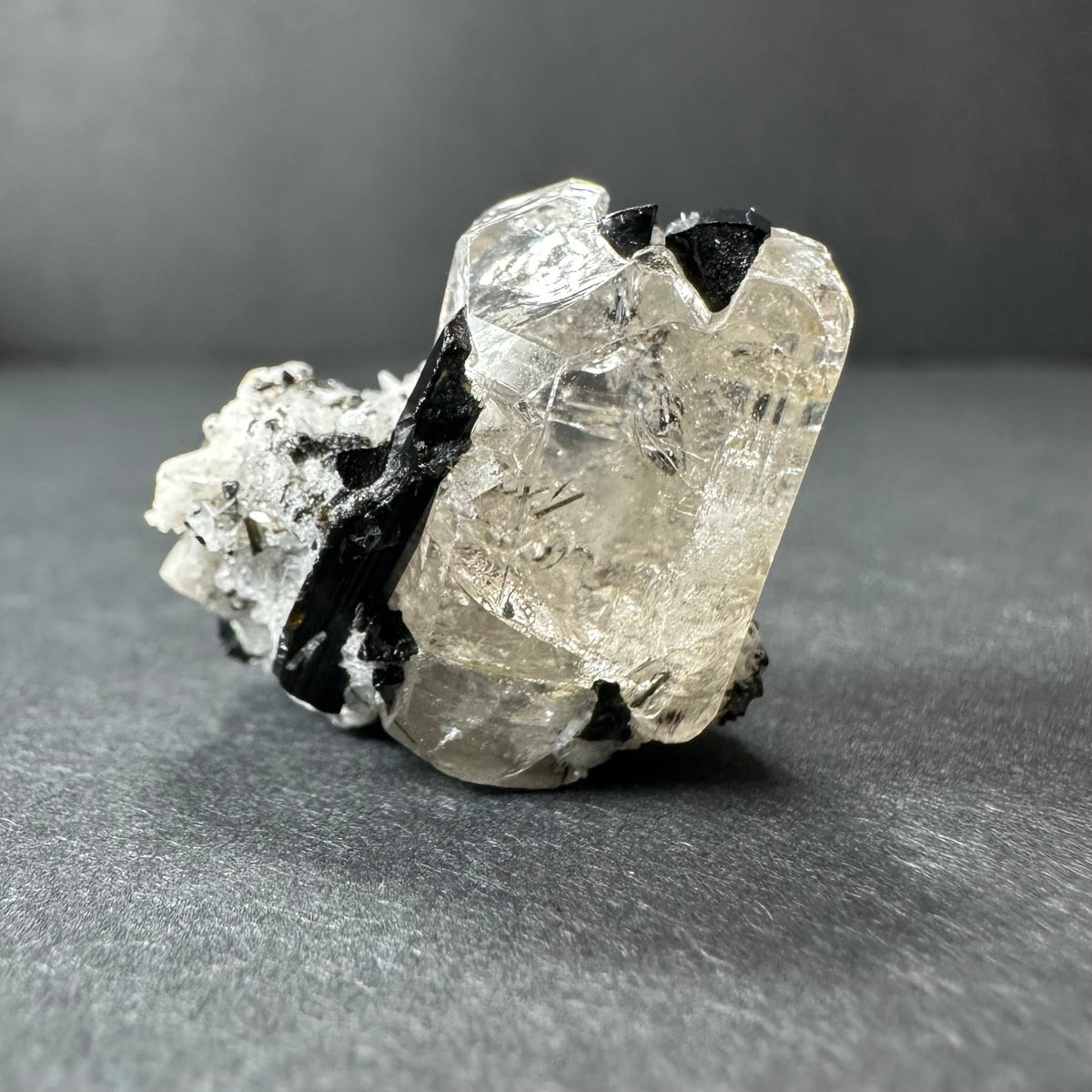 Clear, faceted white topaz crystal with irregular black tourmaline inclusions, resting on a dark gray surface with soft lighting from the left side.