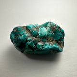 Smooth turquoise nugget with irregular shape and black matrix veins, featuring patches of lighter and darker blue-green hues on a neutral background.