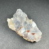 Cluster of translucent pale blue celestite crystals with geometric shapes on a light tan and brown matrix against a dark gray background.