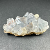 Translucent pale blue celestite crystal cluster with geometric facets resting on a beige matrix, displayed against a dark gray textured background.