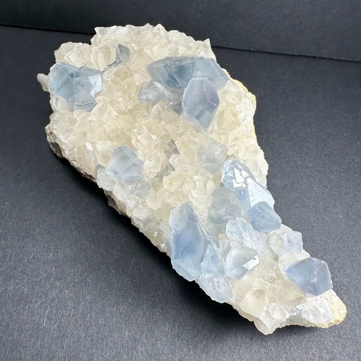 Cluster of translucent pale blue celestite crystals interspersed with white scalenohedral calcite on a rough matrix, displayed against a dark background.
