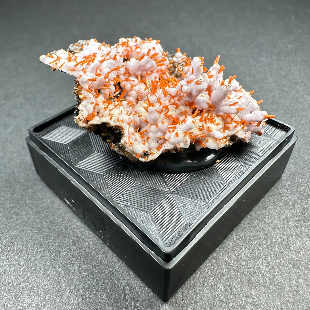 Crocoite crystal cluster with bright orange slender needles emerging from a white, uneven matrix, displayed on a textured black square pedestal against a gray background.