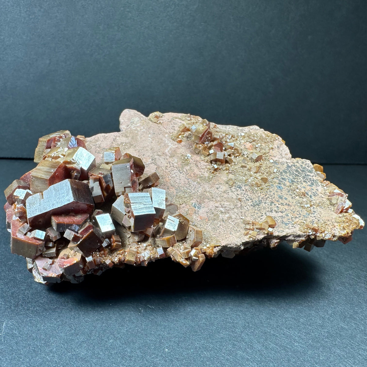 Cluster of reddish-brown vanadinite crystals with shiny hexagonal faces on a light brown matrix, set against a dark background and viewed slightly from the side.