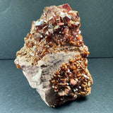 Cluster of deep red to orange vanadinite crystals with hexagonal prism shapes on a rough beige-brown matrix, photographed against a dark background.