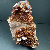 Cluster of reddish-brown vanadinite crystals with hexagonal prisms on a rough beige matrix, shown against a dark background.