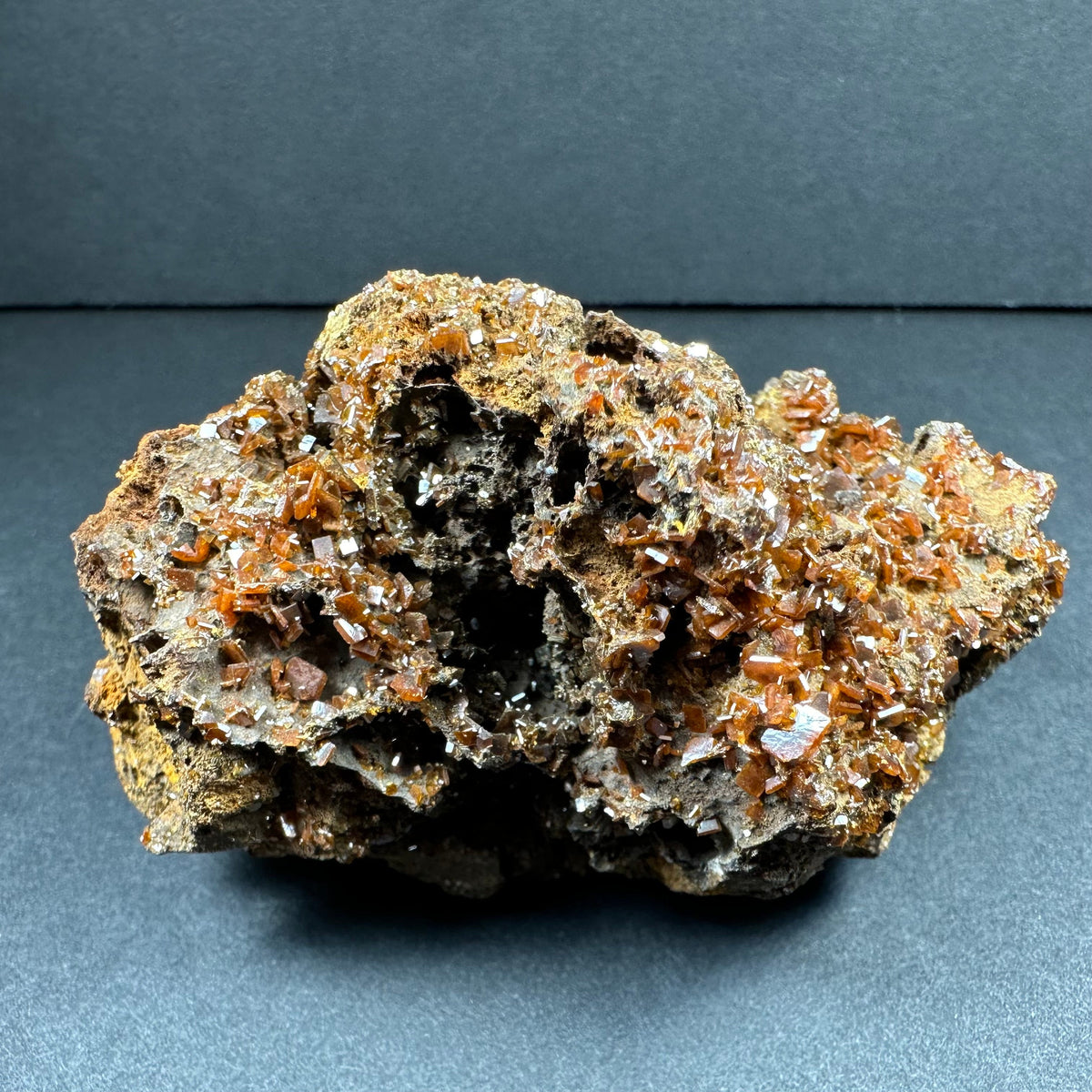 Cluster of orange-brown wulfenite crystals densely covering a rough, gray-brown matrix rock with a hollow central cavity, displayed against a dark background.