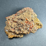 Irregular brownish-yellow rock matrix studded with small, translucent orange-red wulfenite crystals and scattered tiny reflective silver flakes on a dark gray background.