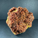 Cluster of reddish-brown vanadinite crystals densely covering an irregular yellowish matrix, photographed against a dark gray background.