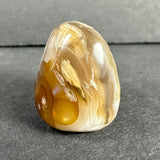 Polished fire agate displaying smooth beige, white, and translucent layers with amber-colored inclusions, set against a textured gray background.