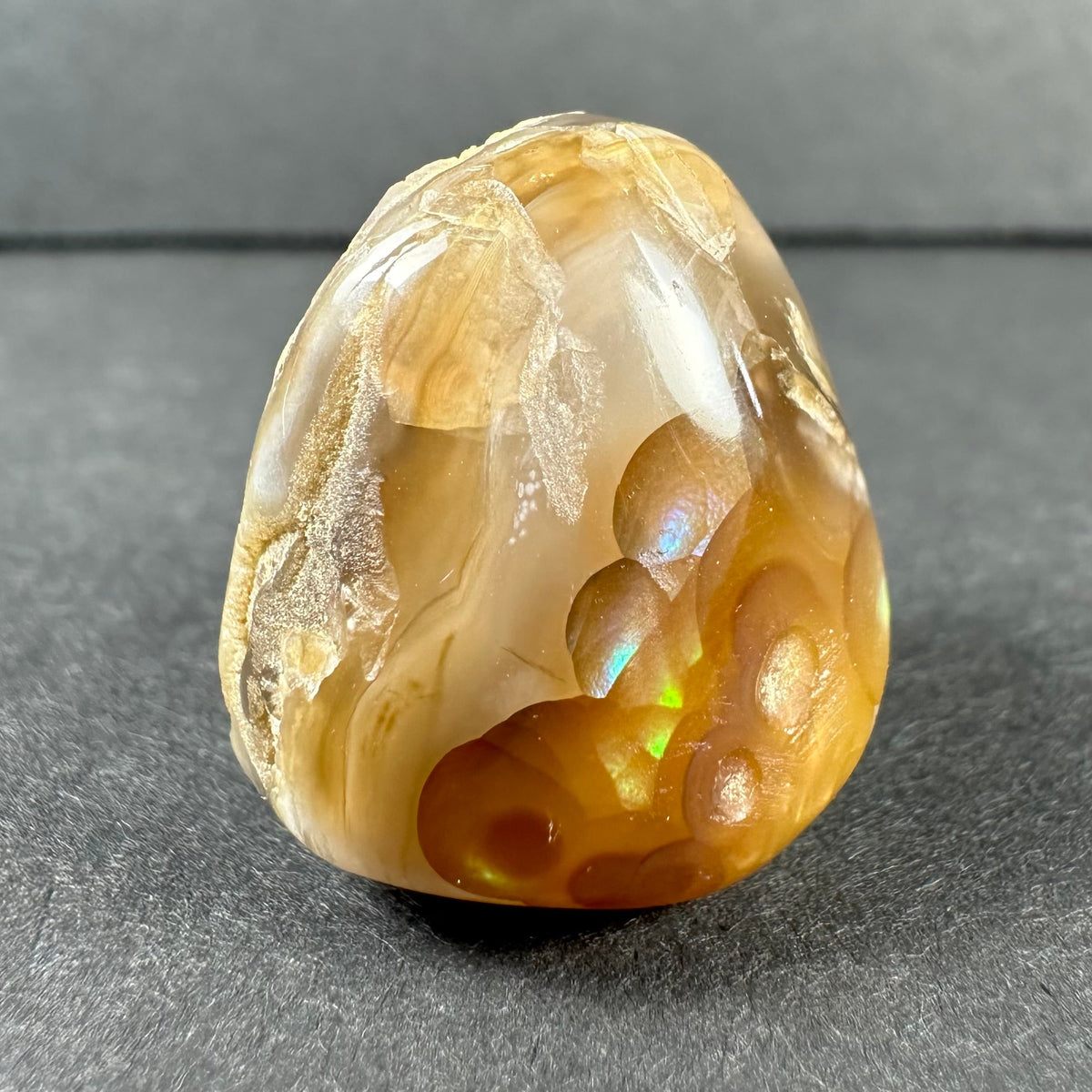 Polished fire agate with amber and cream swirls, featuring iridescent flashes of green and blue on a dark gray background.