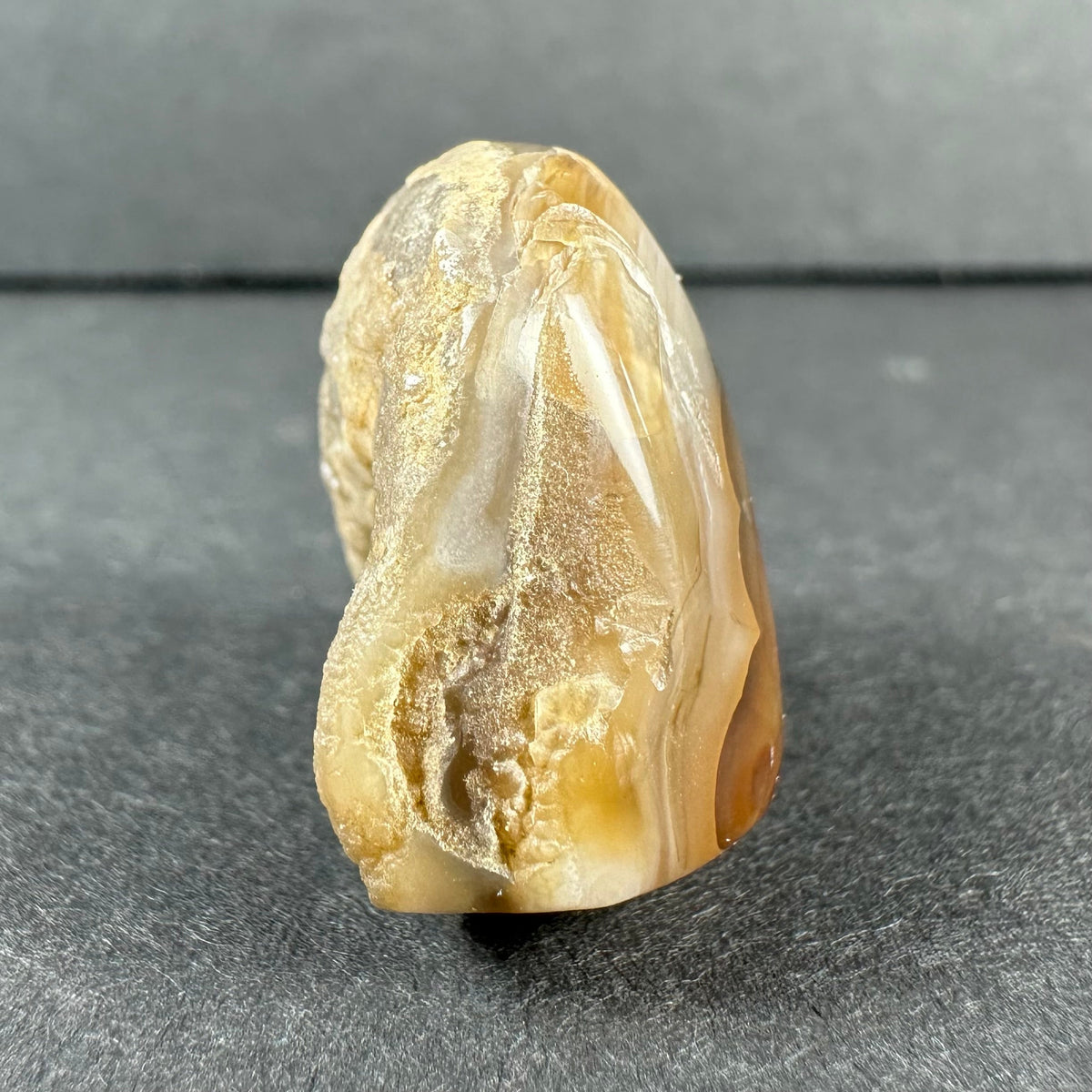 Polished fire agate specimen with layered earthy tones of beige, amber, and cream, showing rough and smooth textures on a dark gray surface.