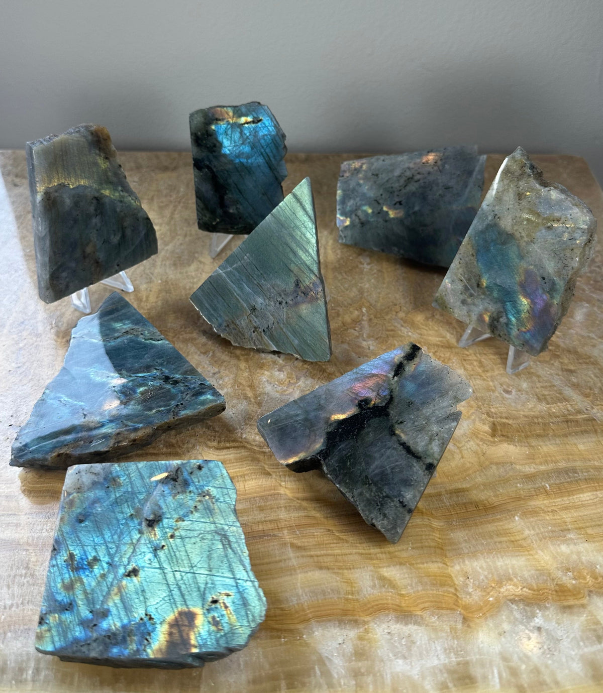Polished labradorite slabs displaying iridescent flashes of blue and green with natural rough gray edges, arranged on a honey-colored wooden surface.