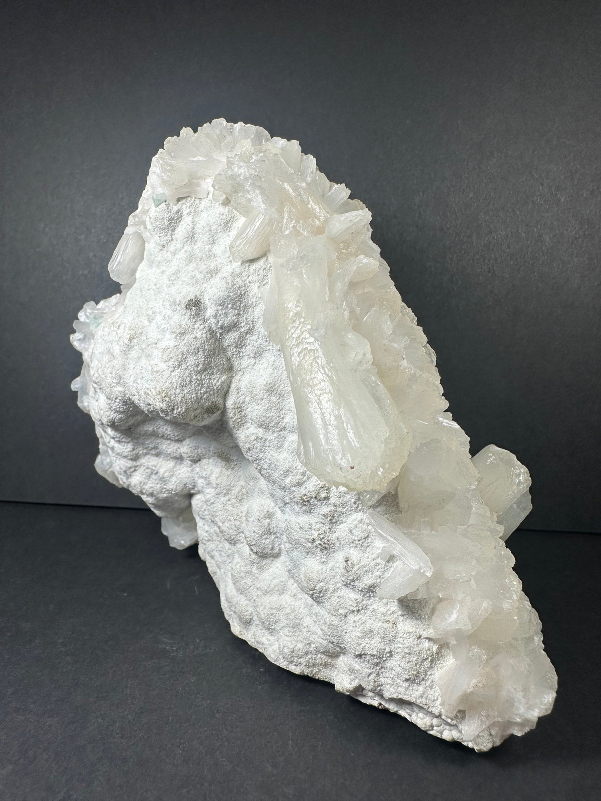 White stilbite crystals clustered on a rough, textured matrix, viewed against a dark gray background highlighting the mineral's natural formation.