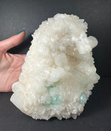 Cluster of white stilbite crystals with small green apophyllite and mordenite formations on a rough gray matrix, held next to a hand for scale