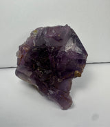 Purple cubic fluorite crystal cluster with translucent to opaque surfaces and sharp geometric edges, resting on a pale matrix against a plain white background.