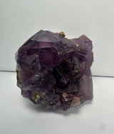 Purple cubic fluorite crystal with translucent, faceted surfaces and natural matrix fragments, set against a plain white background.