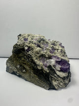 Raw mineral specimen with clusters of purple fluorite crystals and white matrix on a rough, dark rock base shown against a plain gray background.