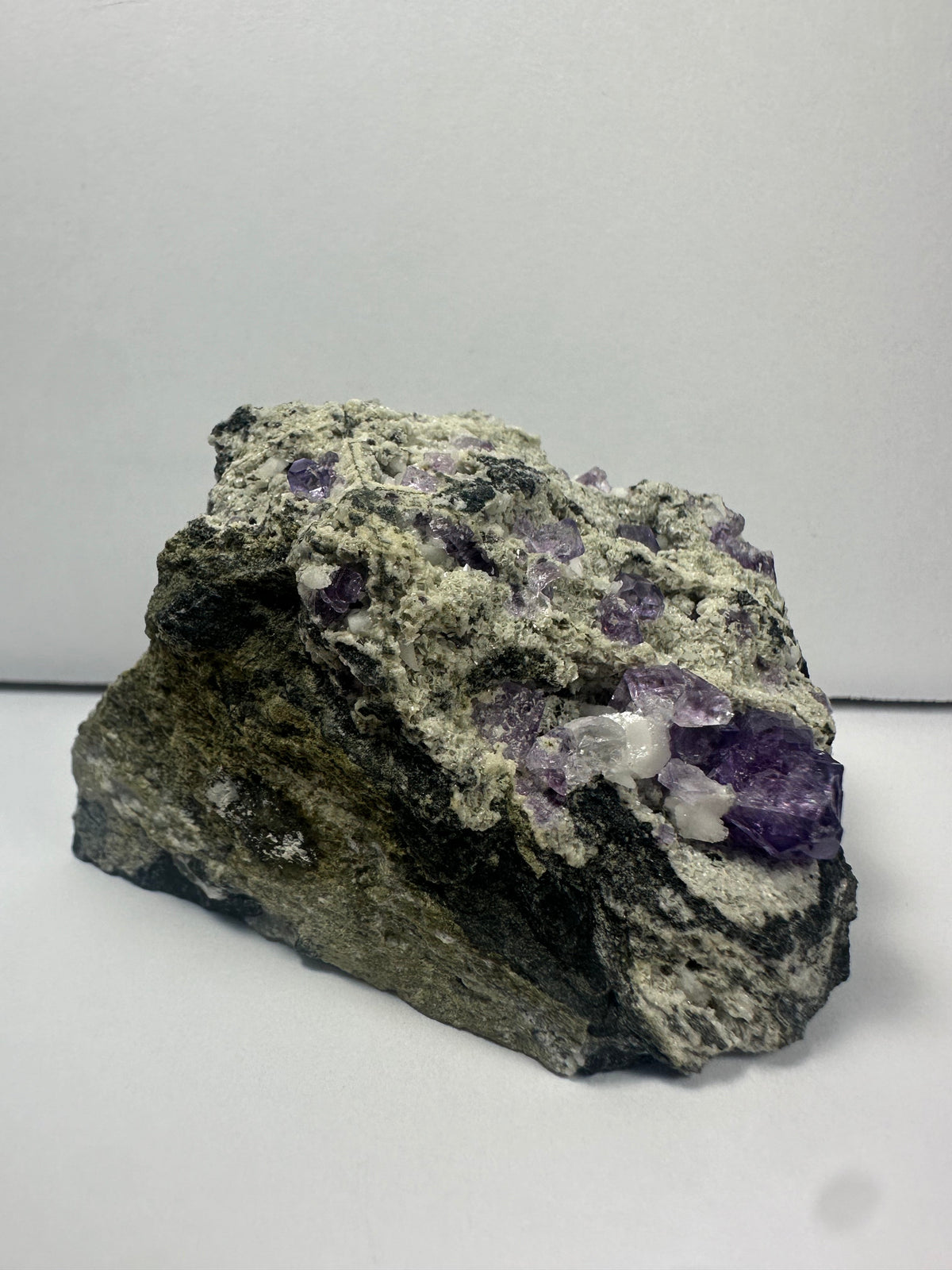Raw mineral specimen with clusters of purple fluorite crystals and white matrix on a rough, dark rock base shown against a plain gray background.