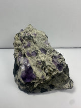 Chunky mineral rock with clusters of deep purple fluorite crystals embedded in a rough grayish-green matrix, viewed against a plain light background.
