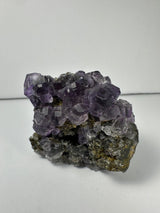 Cluster of purple dodecahedral fluorite crystals with calcite on a dark mineral matrix, displaying translucent and geometric crystal shapes.