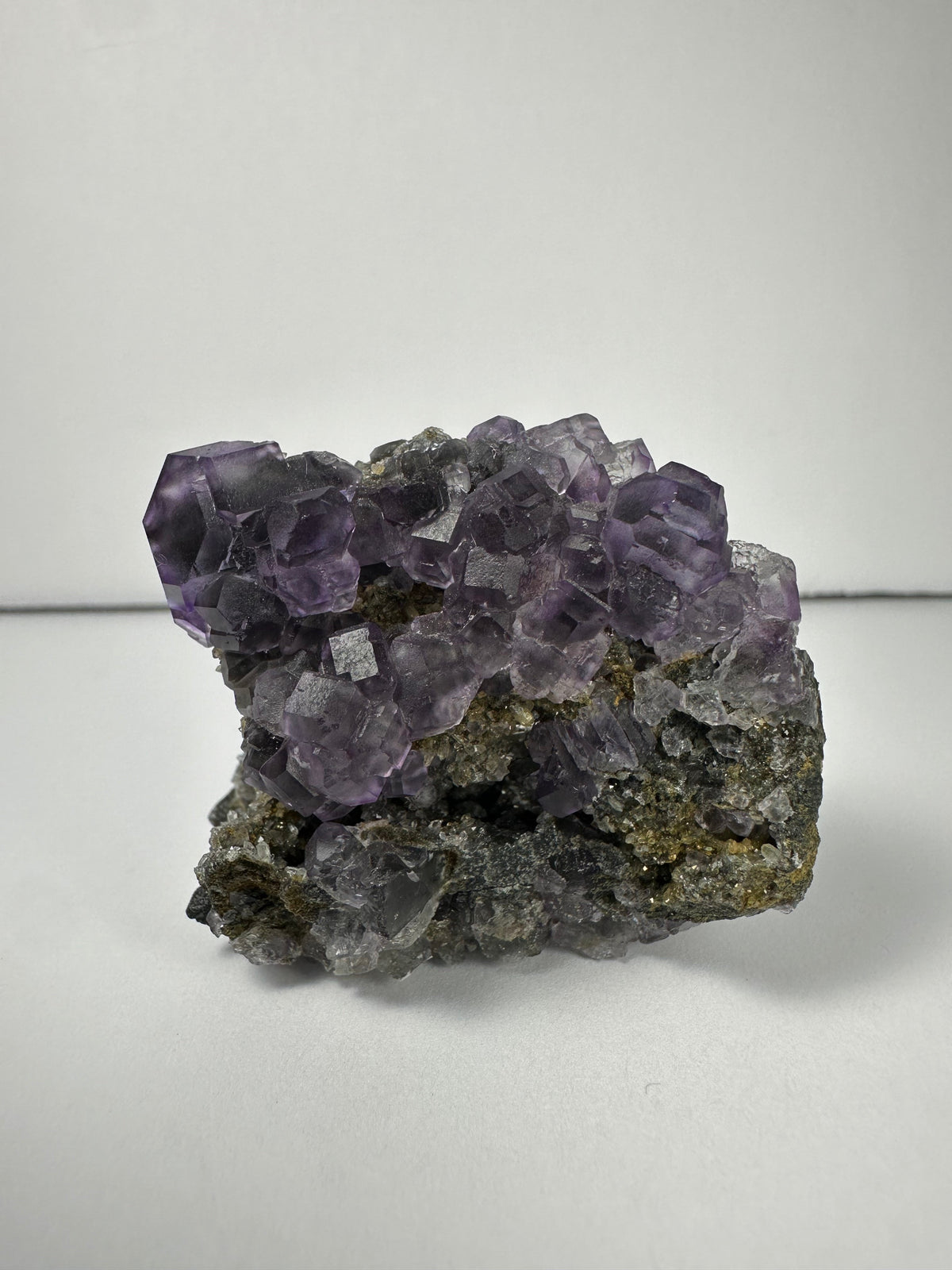 Cluster of translucent purple dodecahedral fluorite crystals atop a gray and brown matrix, showing well-defined geometric shapes and slight surface texture variations.