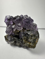 Cluster of translucent purple dodecahedral fluorite crystals atop a rough, dark mineral matrix with small white calcite crystals, shown against a plain light background.