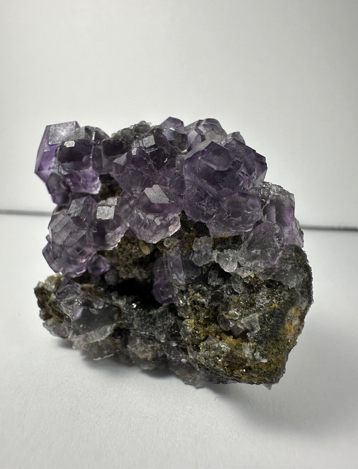 Cluster of translucent purple dodecahedral fluorite crystals atop a rough, dark mineral matrix with small white calcite crystals, shown against a plain light background.