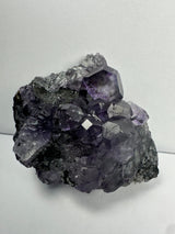 Cluster of purple dodecahedral fluorite crystals with translucent calcite formations on a dark matrix, displayed against a plain white background.