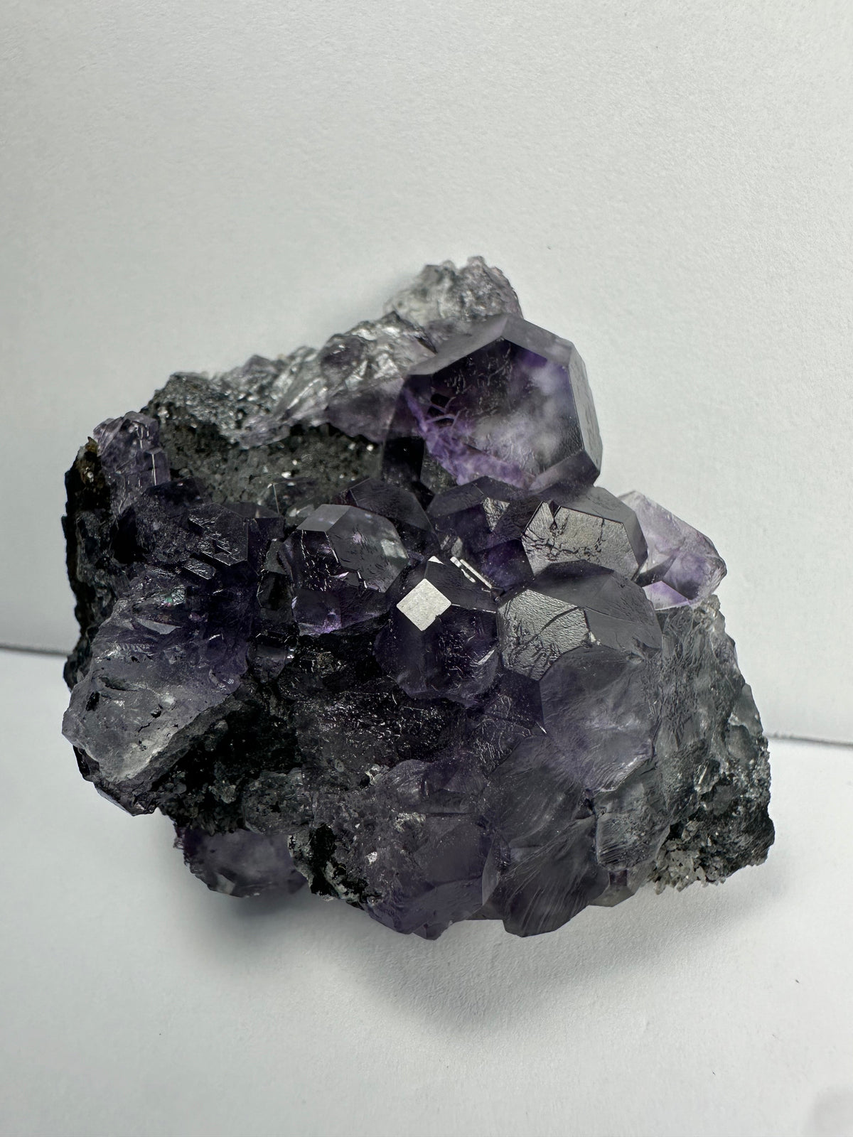 Cluster of purple dodecahedral fluorite crystals with translucent calcite formations on a dark matrix, displayed against a plain white background.