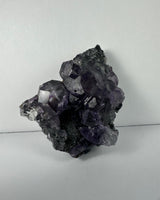 Cluster of dark purple dodecahedral fluorite crystals with some white calcite on a rough gray matrix, shown against a plain light background.