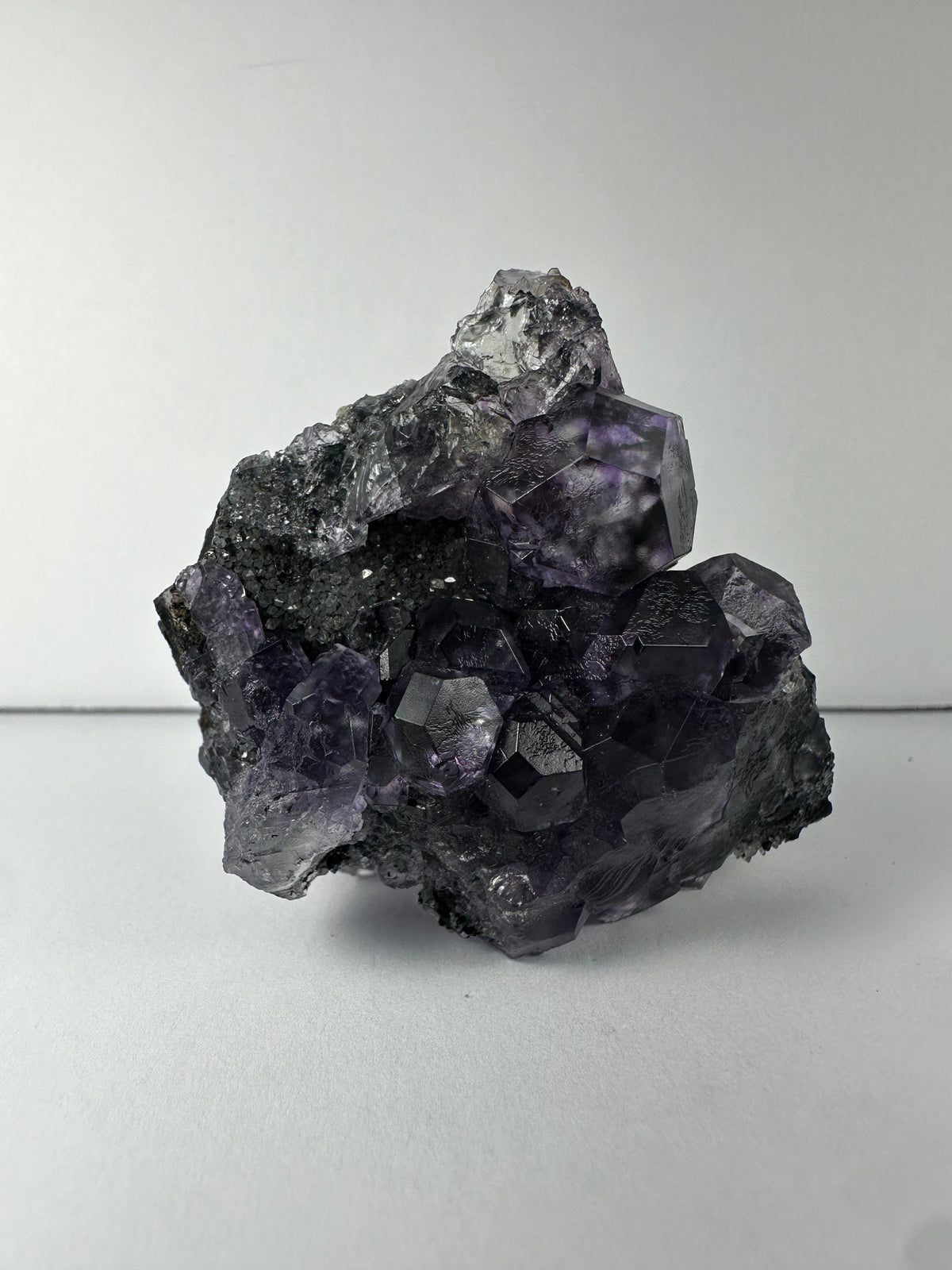 Cluster of translucent purple dodecahedral fluorite crystals with a rough gray calcite matrix, showing geometric facets and layered textures.