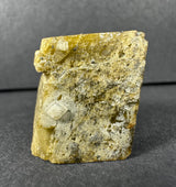 Yellow-tan rough mineral block with small white diamond quartz crystals on its textured surface, set against a dark gray background.