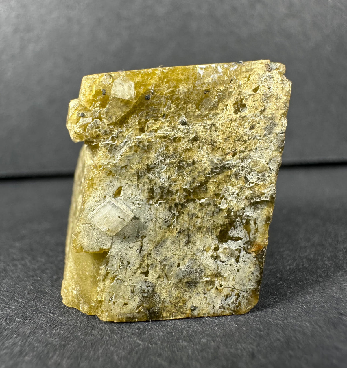 Yellow-tan rough mineral block with small white diamond quartz crystals on its textured surface, set against a dark gray background.