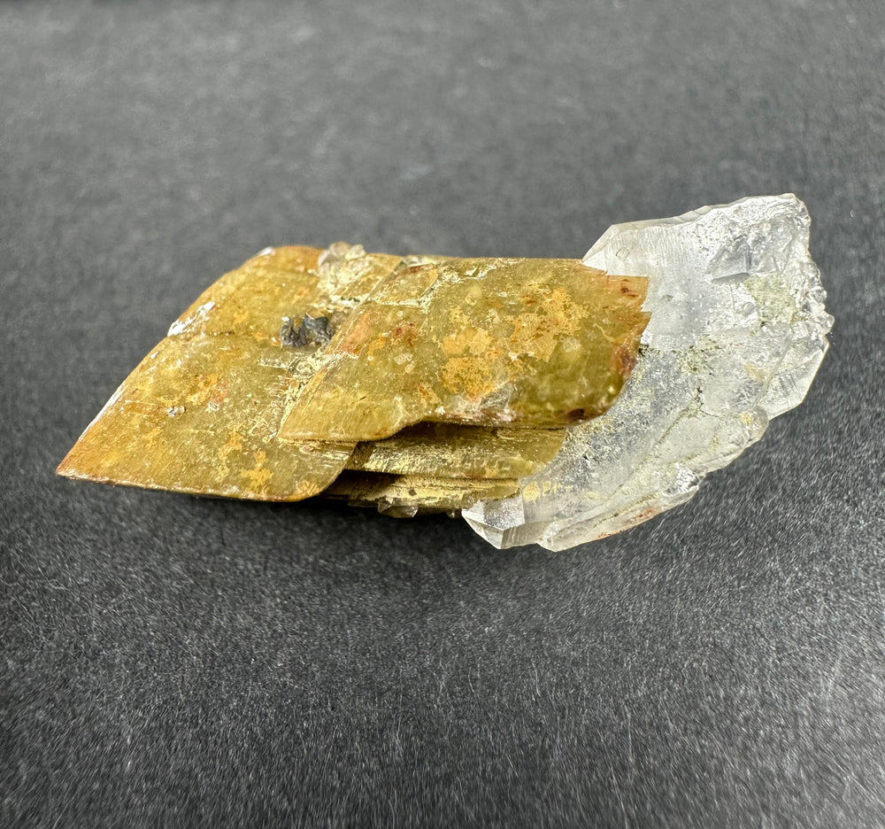 Transparent diamond quartz crystal partially attached to layered golden-brown siderite mineral base, displayed on a dark gray textured surface.