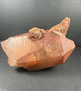 Tangerine quartz crystal cluster with reddish-orange hues and rough, textured formations from the Atlas Mountains, Morocco, against a neutral dark background.