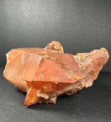 Orange-tinted Tangerine quartz cluster with sharp crystalline faces and rough textured base against a dark background.