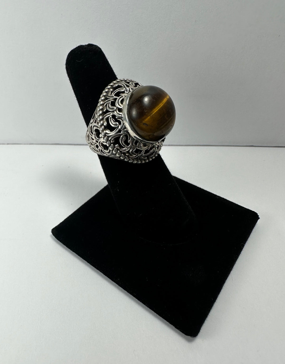 Ornate silver ring with a large polished tiger's eye stone, displayed on a black velvet stand against a plain white background.