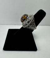 Silver ring with intricate filigree design featuring a polished dome-shaped tiger's eye stone, displayed upright on a black velvet stand.